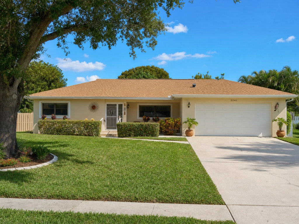 A tan single-story home in Greenacres featuring a clean brown architectural shingle roof and a large shade tree in the front yard.
